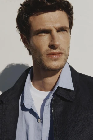 Portrait of a handsome adult male model in a blue collared shirt and black jacket against a white background in natural light.