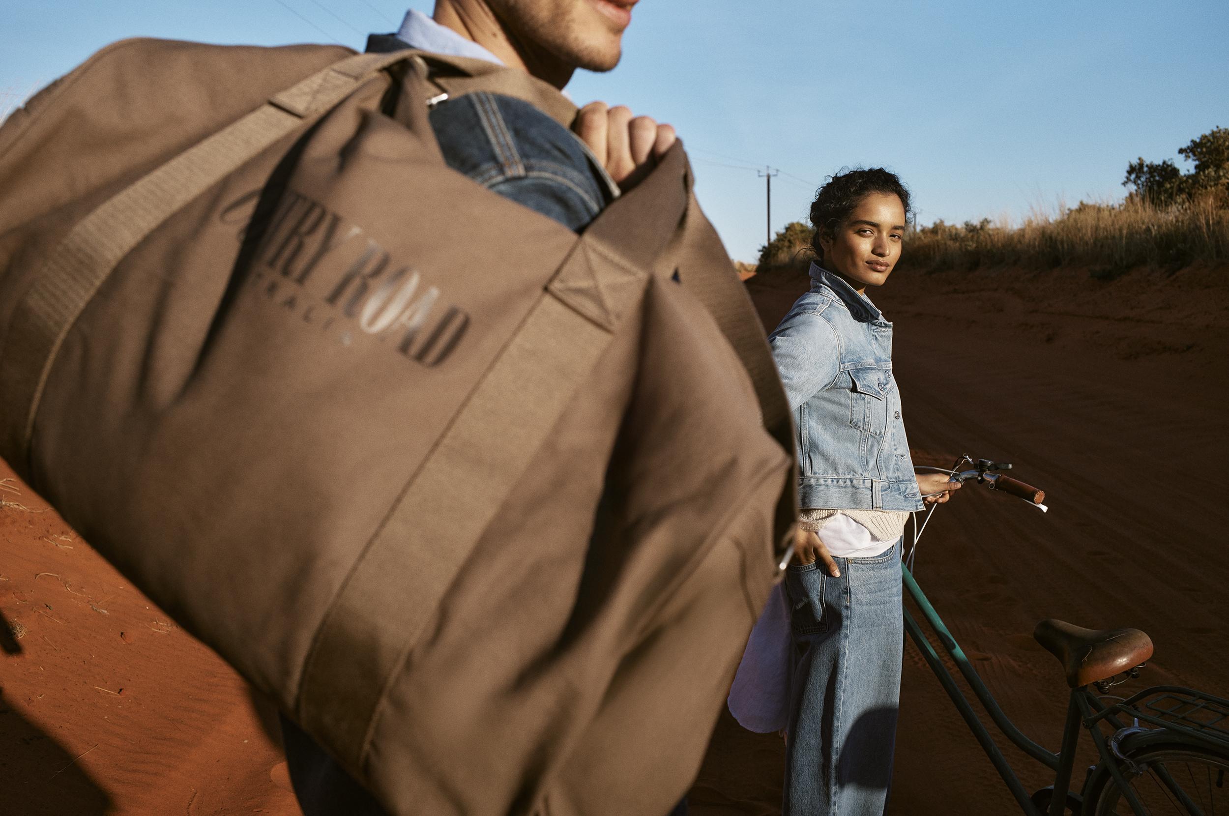 Two people in a dry, rural outdoor setting. A person carries a brown Country Road duffel bag, with another person and a bicycle nearby.