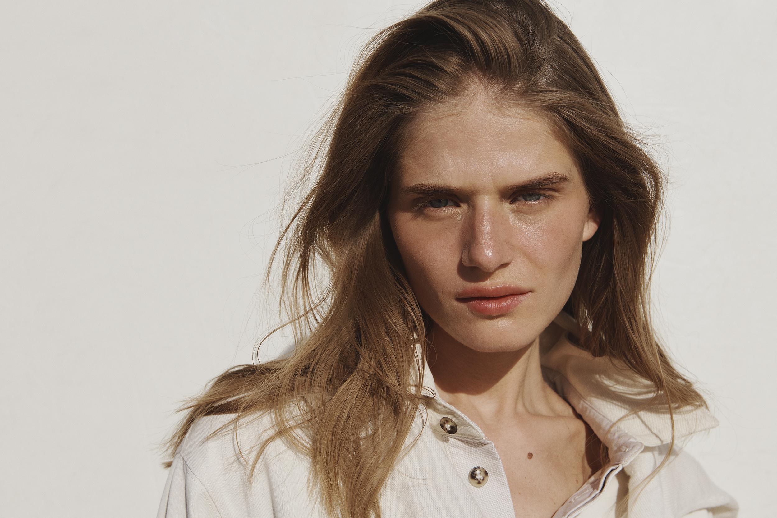 Close-up studio portrait of a young adult woman with long brown hair, wearing a button-down shirt against a bright white background.