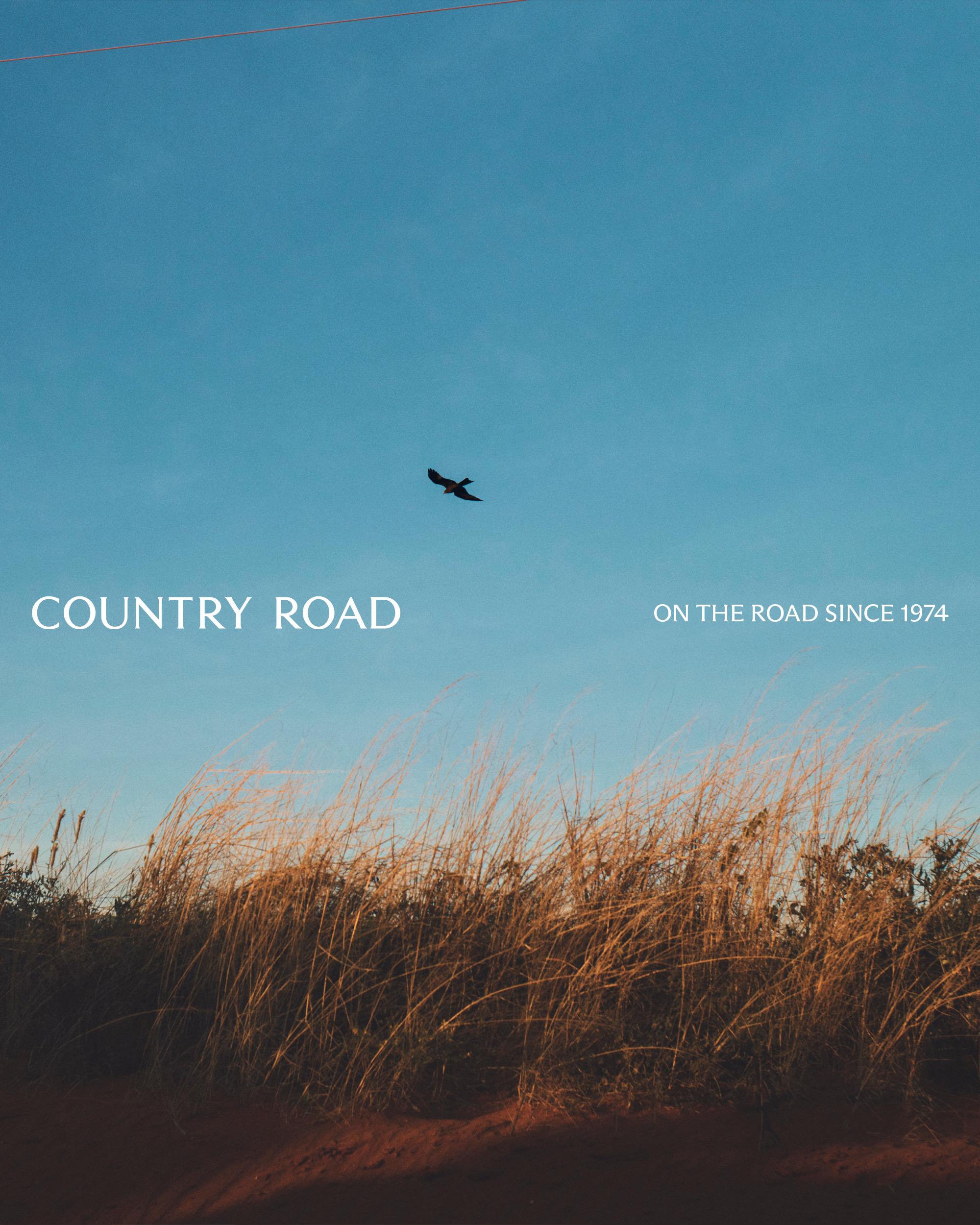 A bird flies over tall grass against a blue sky with the words 'Country Road. On the road since 1974' displayed on the upper left.