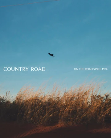 A bird flies over tall grass against a blue sky with the words 'Country Road. On the road since 1974' displayed on the upper left.