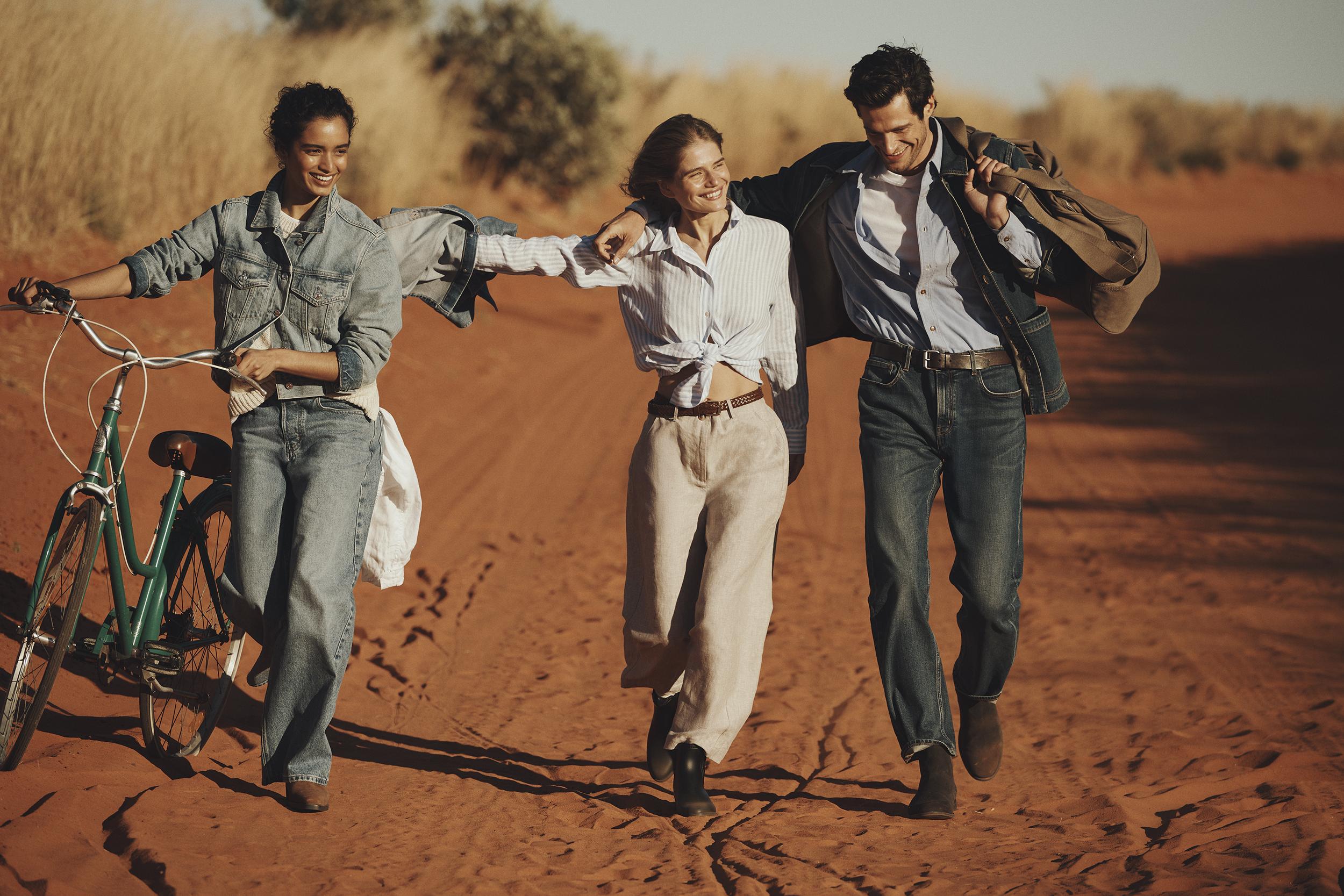 Three people walk and push a bicycle on a red dirt road in a desert landscape. Denim and casual clothing in a sunlit scene.