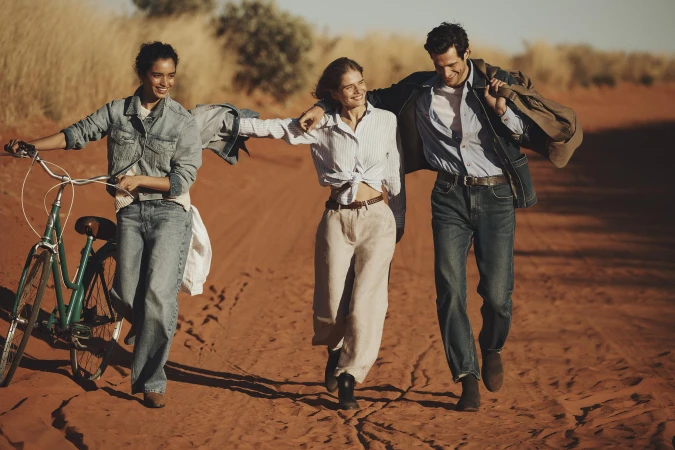 Three people walk and push a bicycle on a red dirt road in a desert landscape. Denim and casual clothing in a sunlit scene.
