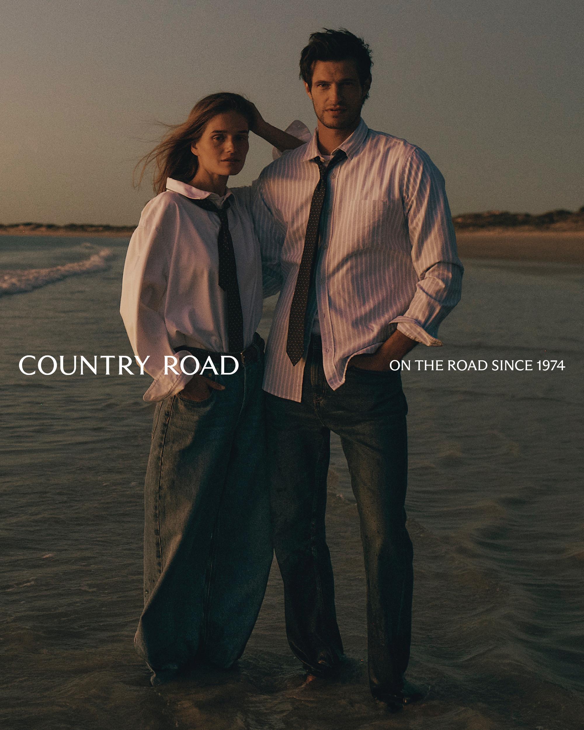 Fashion shot of a man and woman standing on a beach near the water, both wearing white shirts and dark ties with a relaxed pose.