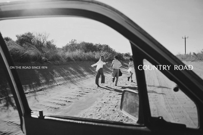 Three people are seen running down a dirt road in a black and white image, as viewed through the open window of a car.
