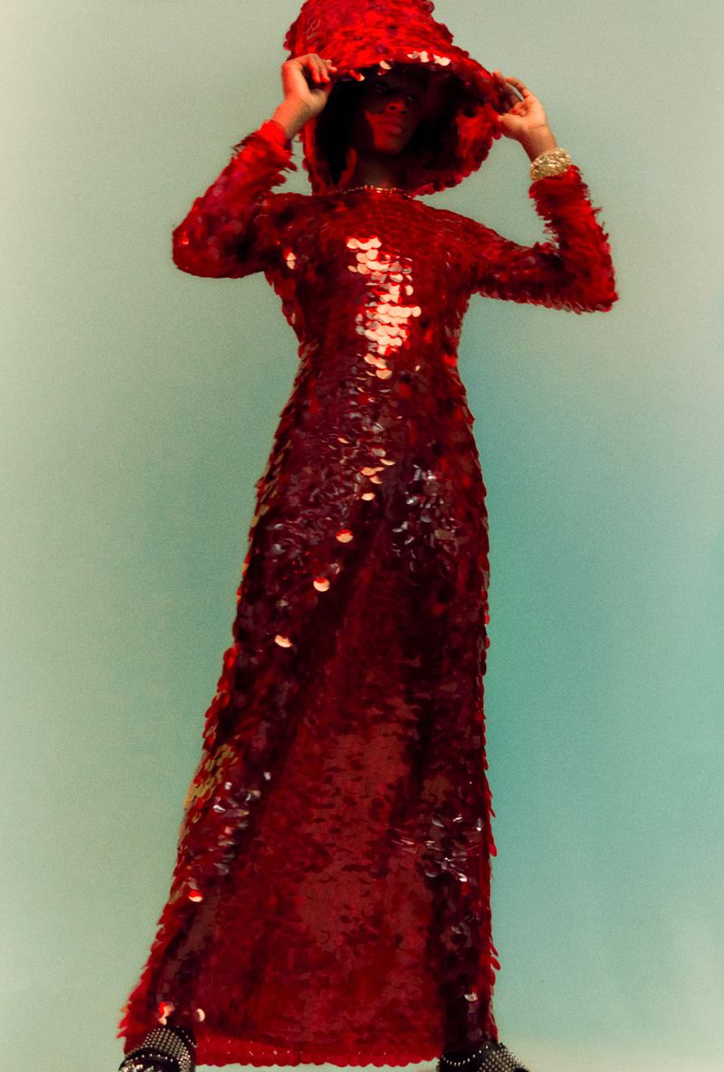 A woman stands in a red sequined dress with a hood and platform shoes against an aqua background in a studio fashion shot.