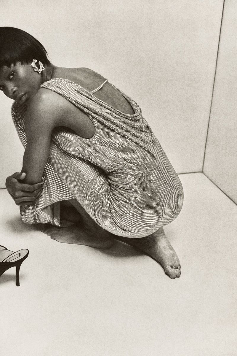 Monochromatic fashion shot of a woman in a glittering dress, crouching indoors with high-heeled shoes nearby against a neutral backdrop.