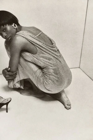 Monochromatic fashion shot of a woman in a glittering dress, crouching indoors with high-heeled shoes nearby against a neutral backdrop.