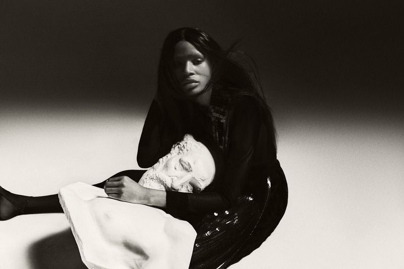 Monochrome fashion shot features a Black woman embracing a Greco-Roman sculpture head, set against a softly lit background, evoking fine art.