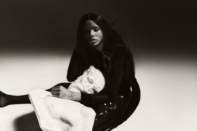 Monochrome fashion shot features a Black woman embracing a Greco-Roman sculpture head, set against a softly lit background, evoking fine art.
