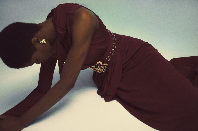 An adult woman with short hair wearing a maroon dress poses in front of a white background in a studio environment, accessorized with jewelry.