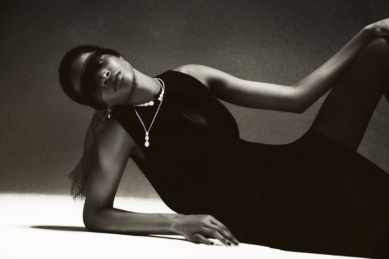 Adult female model in black dress laying down wearing pearl jewelry and sheer black fabric over the eyes in a photo studio.