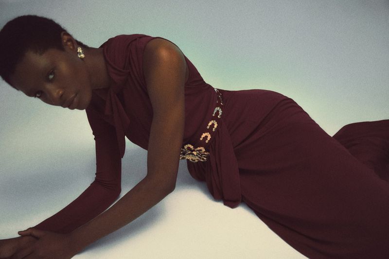 Model Onyinyechi Chijioke reclined in a burgundy long-sleeved dress with an embellished belt against a pale studio backdrop.