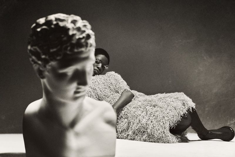 Monochromatic studio portrait of Black woman draped in textured fabric, reclining next to a classical sculpture bust against a dark backdrop.