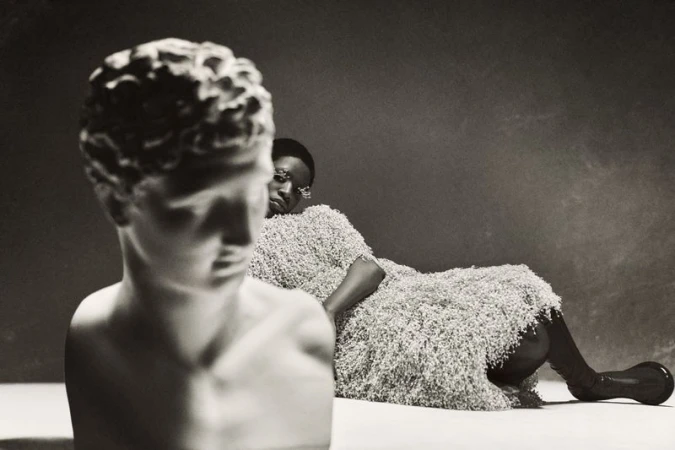 Monochromatic studio portrait of Black woman draped in textured fabric, reclining next to a classical sculpture bust against a dark backdrop.