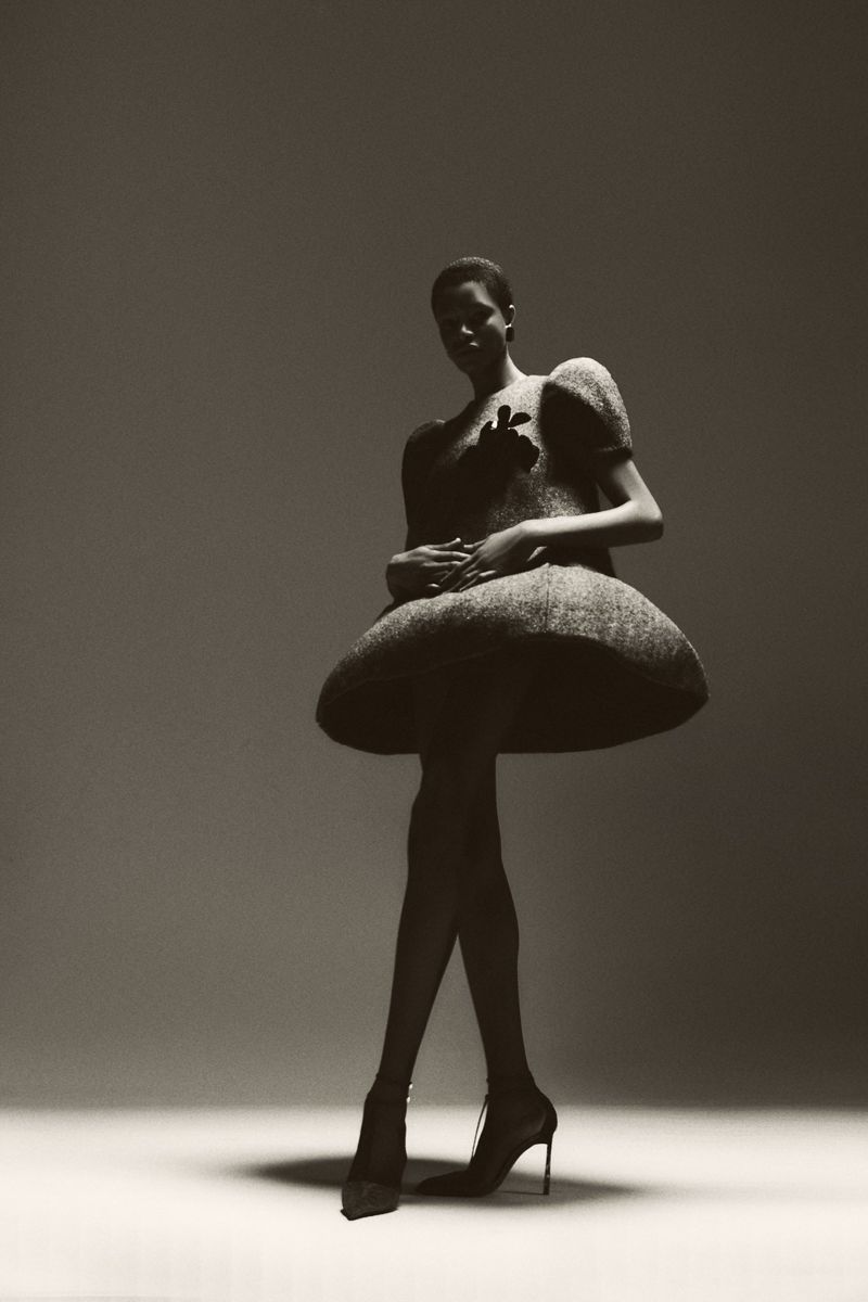 Full-length shot of a Black fashion model in a sculptural dress and high heels, standing in a photo studio with a grey background and dramatic lighting.