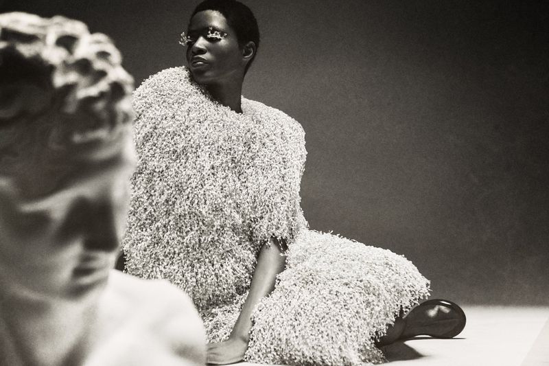 Artistic studio shot of a woman in a furry dress seated next to a sculpture, creating a black and white fashion portrait.