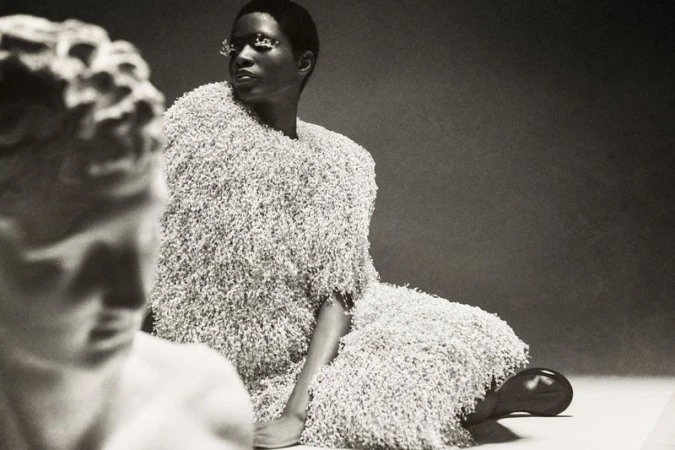 Artistic studio shot of a woman in a furry dress seated next to a sculpture, creating a black and white fashion portrait.