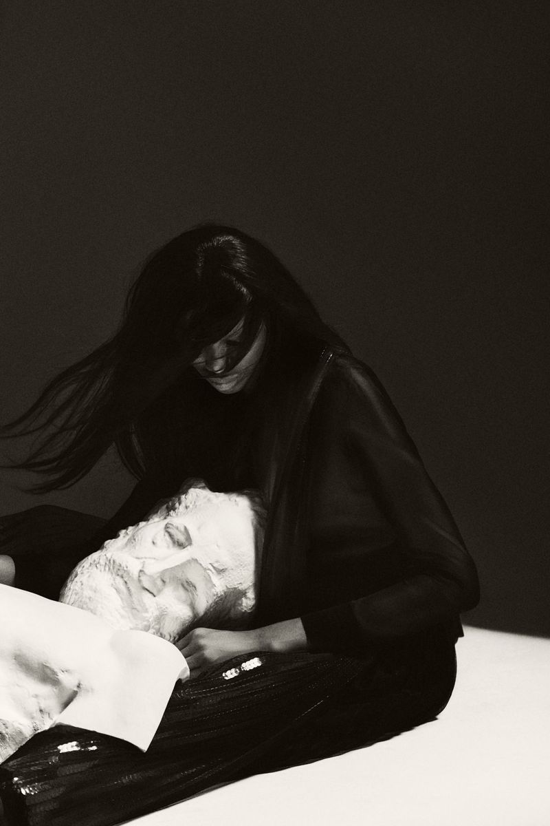 Woman in black leans over a man lying down, both in monochrome. Studio lighting emphasizes their forms and clothing.