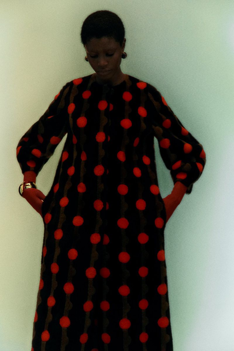 Elegant Black woman in a retro-style black and red polka dot dress indoors, accessorized with a watch and statement earrings.