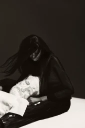Model Onyinyechi Chijioke seated in dark clothing leaning over a white classical sculpted bust in low-key lighting.