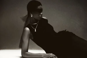 Model Onyinyechi Chijioke reclines in a black sleeveless dress and mesh eye veil with drop jewelry in a dark studio setting.