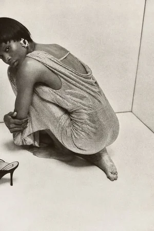 Model Onyinyechi Chijioke crouched barefoot in a corner wearing a draped metallic mesh open-back dress.