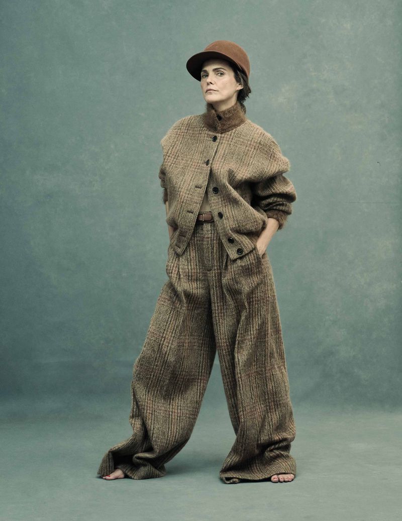 Woman in tweed suit with hat poses in front of textured green backdrop in a studio. Fashion editorial photography.