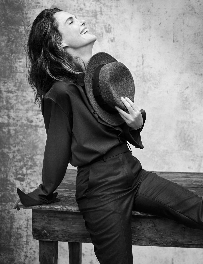 Monochrome portrait of smiling woman in dark blouse and pants, leaning on wooden table, holding hat against mottled background.