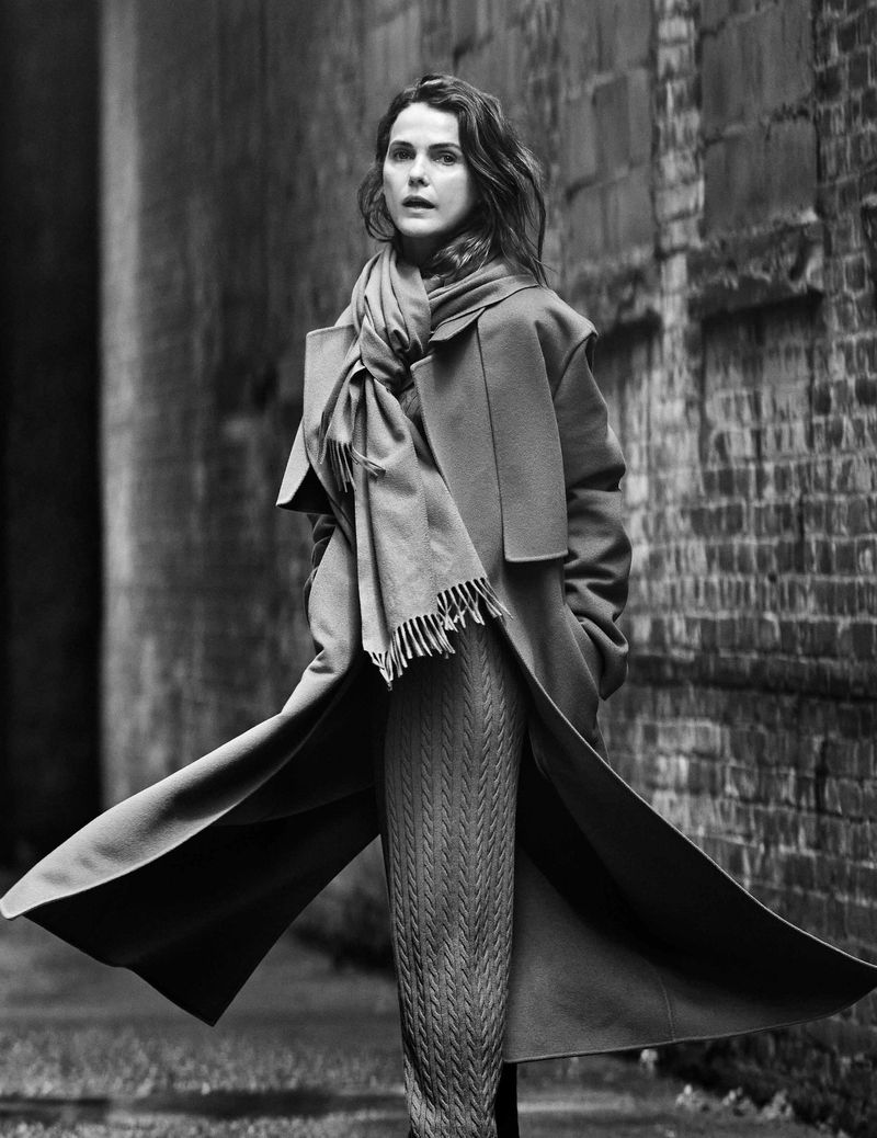 Monochrome portrait of a stylish adult woman in a long coat and scarf, standing against a weathered brick wall in an outdoor alleyway.