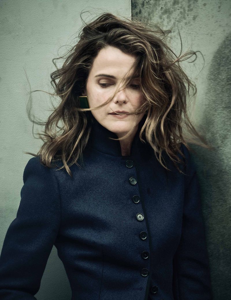 Portrait of a woman with wavy hair, wearing a dark buttoned jacket, against a textured gray backdrop. Soft, diffused lighting.