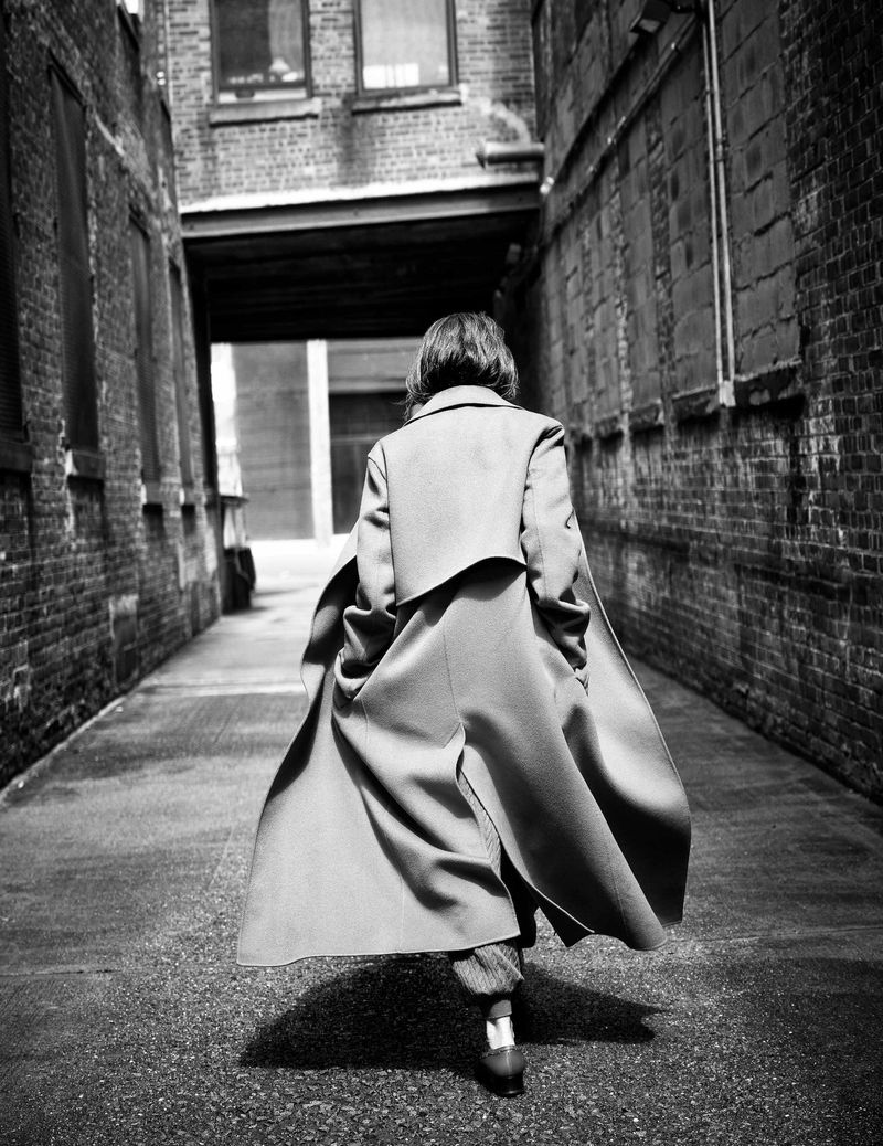 Person in long coat walks down a narrow alleyway with aged brick walls. Black and white fashion photography with urban texture.