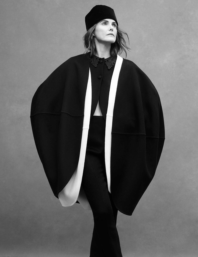 Fashionable adult female in a black and white cloak and hat poses against a grey background in a studio fashion shot.
