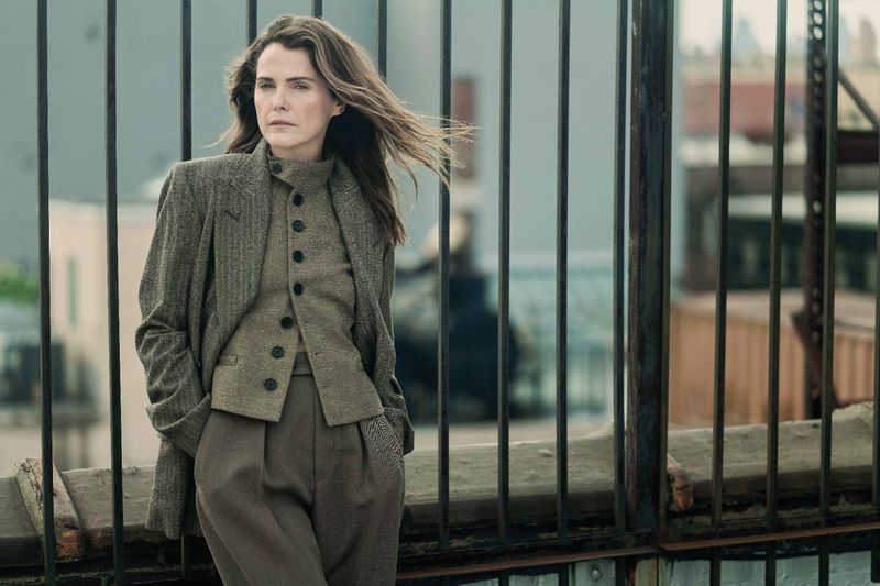 A woman in a wool suit stands outside with a dark metal fence in the foreground and buildings in the distance, showcasing fashion.