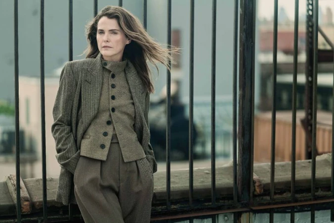 A woman in a wool suit stands outside with a dark metal fence in the foreground and buildings in the distance, showcasing fashion.