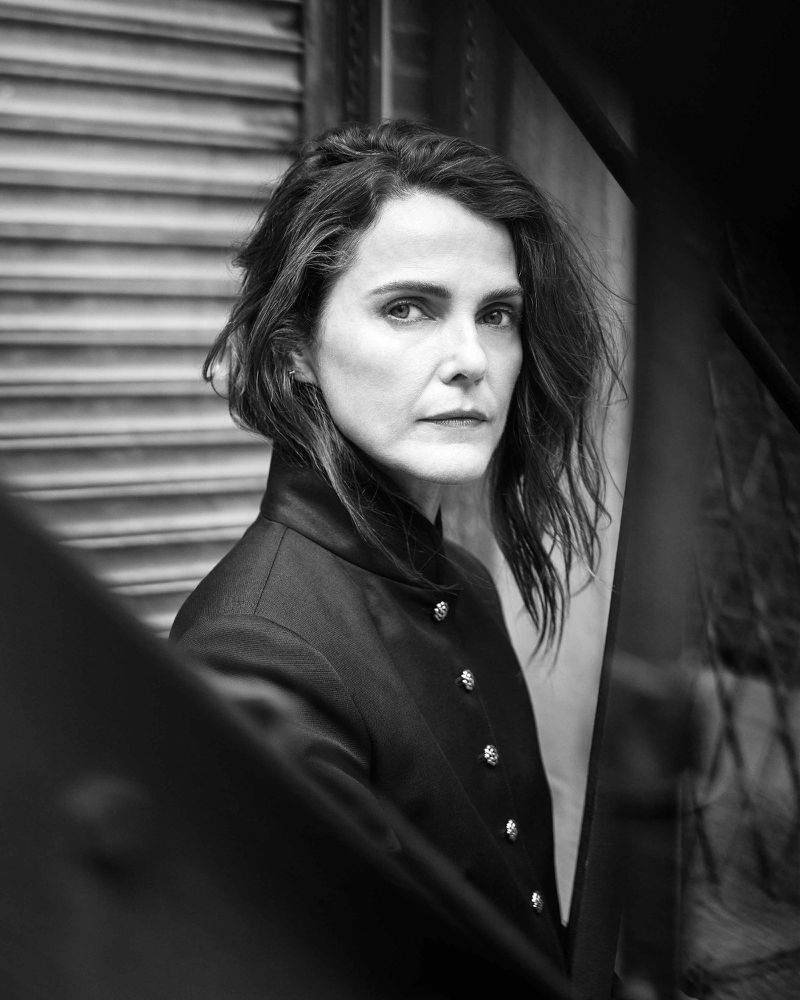 Black and white portrait of a mature woman with short wavy hair wearing a coat in front of a corrugated metal wall.