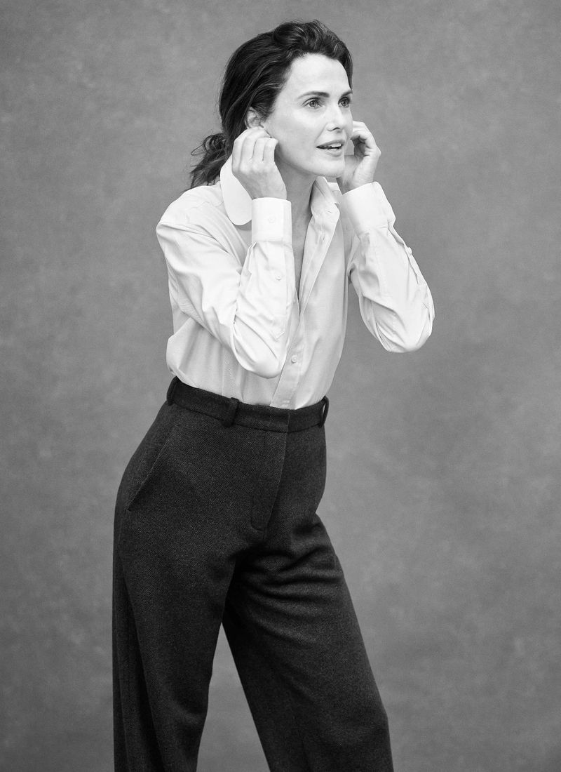Black and white portrait of an adult woman in a white shirt and dark trousers, looking off to the side with her hands near her ears.