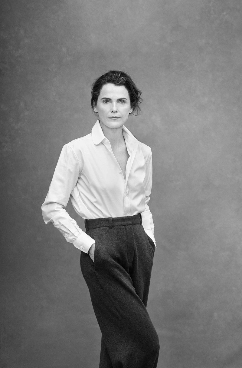 Black-and-white portrait of a woman with dark hair in a white shirt and dark pants with her hands in her pockets against a gray background.