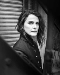 Black and white portrait of Keri Russell wearing a black high-collar buttoned coat framed by architectural metalwork.