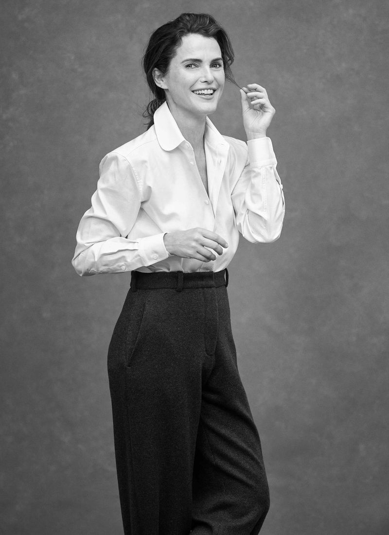 Black and white studio portrait of Keri Russell smiling in a white button-down shirt and high-waisted dark trousers.