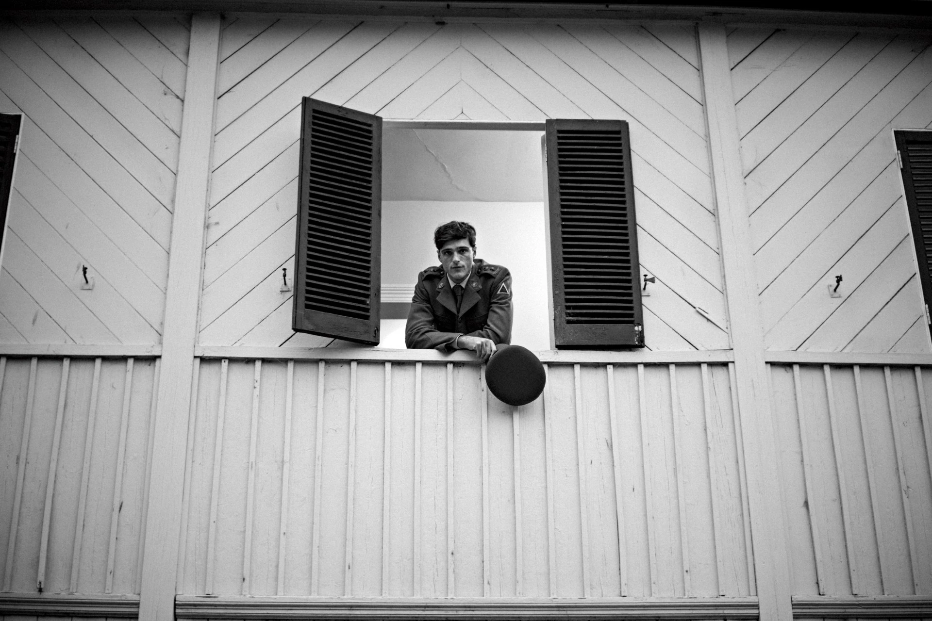 Monochrome portrait of a man in military attire leaning on a window, holding his hat in hand, with a somber expression. Black and white.