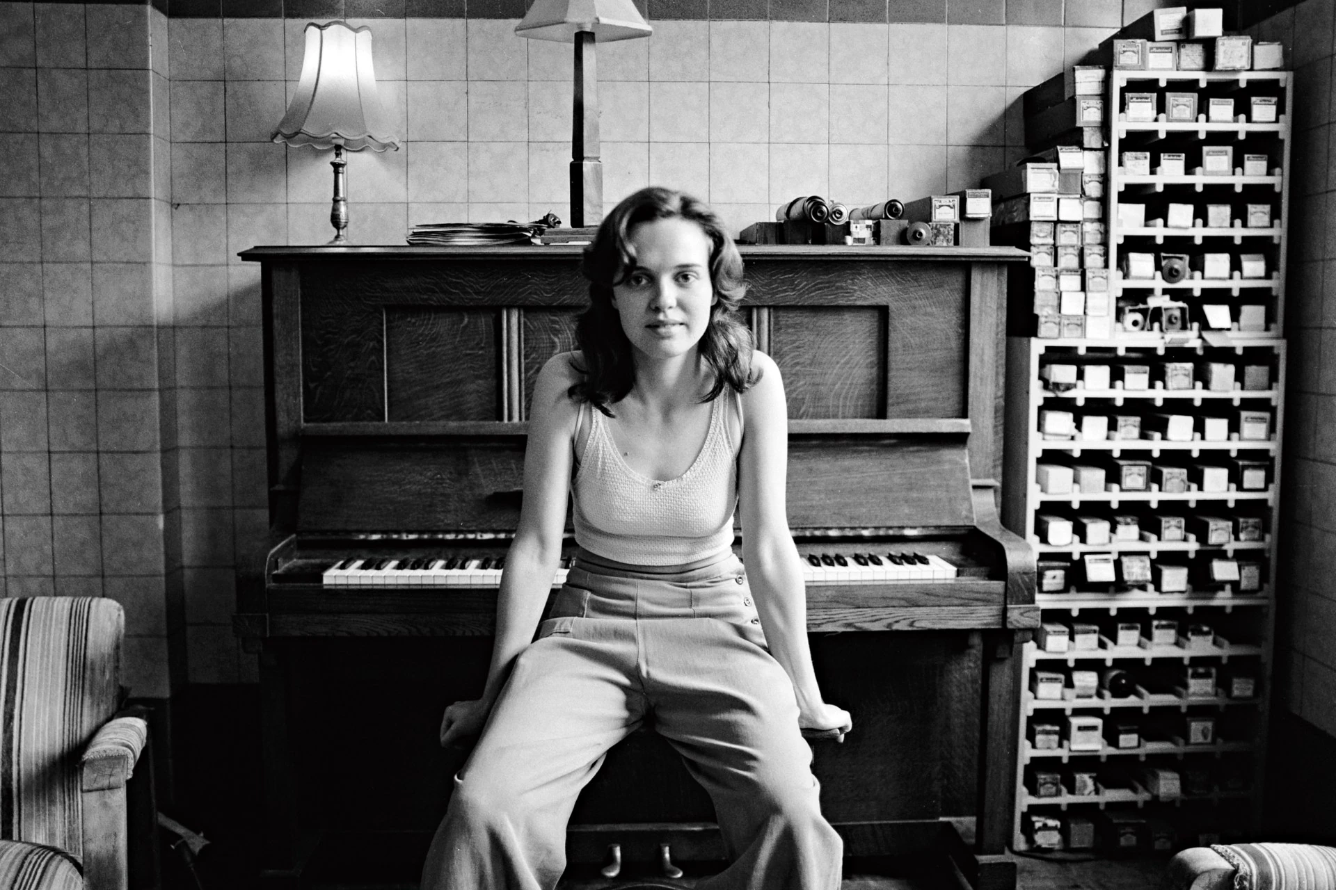 Woman sitting on a piano bench in front of an upright piano, indoor setting with tiled walls, vintage aesthetic, black and white photography.
