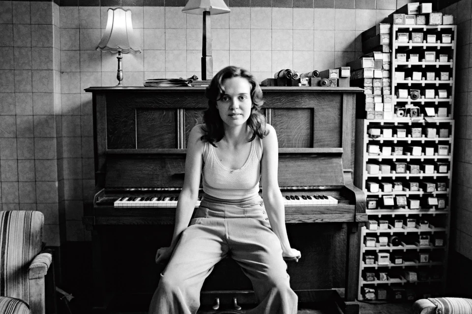 Woman sitting on a piano bench in front of an upright piano, indoor setting with tiled walls, vintage aesthetic, black and white photography.