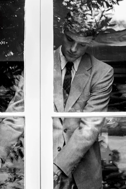 Monochrome portrait of a man in a suit looking down through a window, with a blurred outdoor background, creating an introspective mood.
