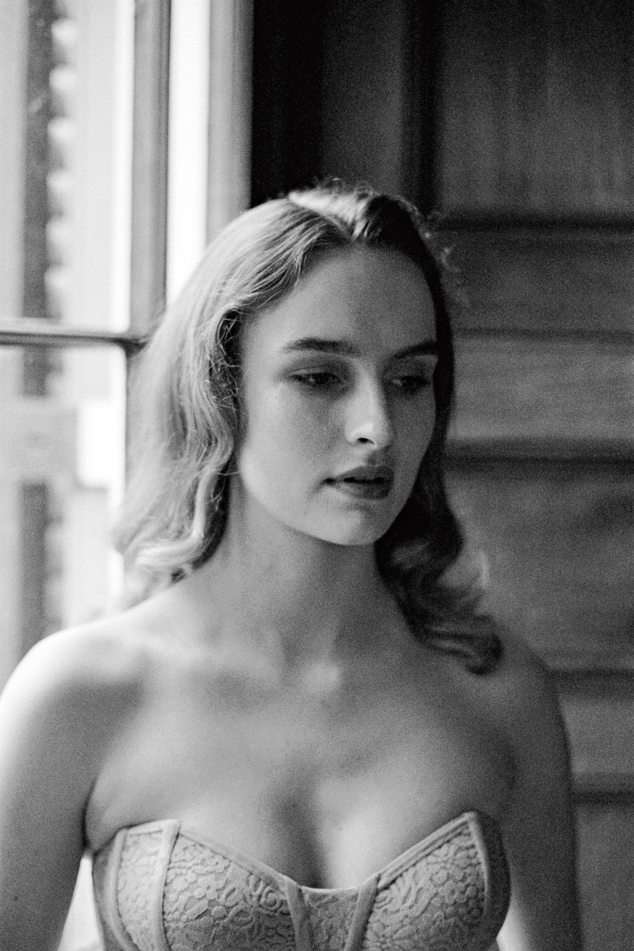 Black and white portrait of a woman in a corset near a window with natural light, showcasing soft curls and indoor fashion photography.