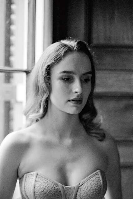 Black and white portrait of a woman in a corset near a window with natural light, showcasing soft curls and indoor fashion photography.