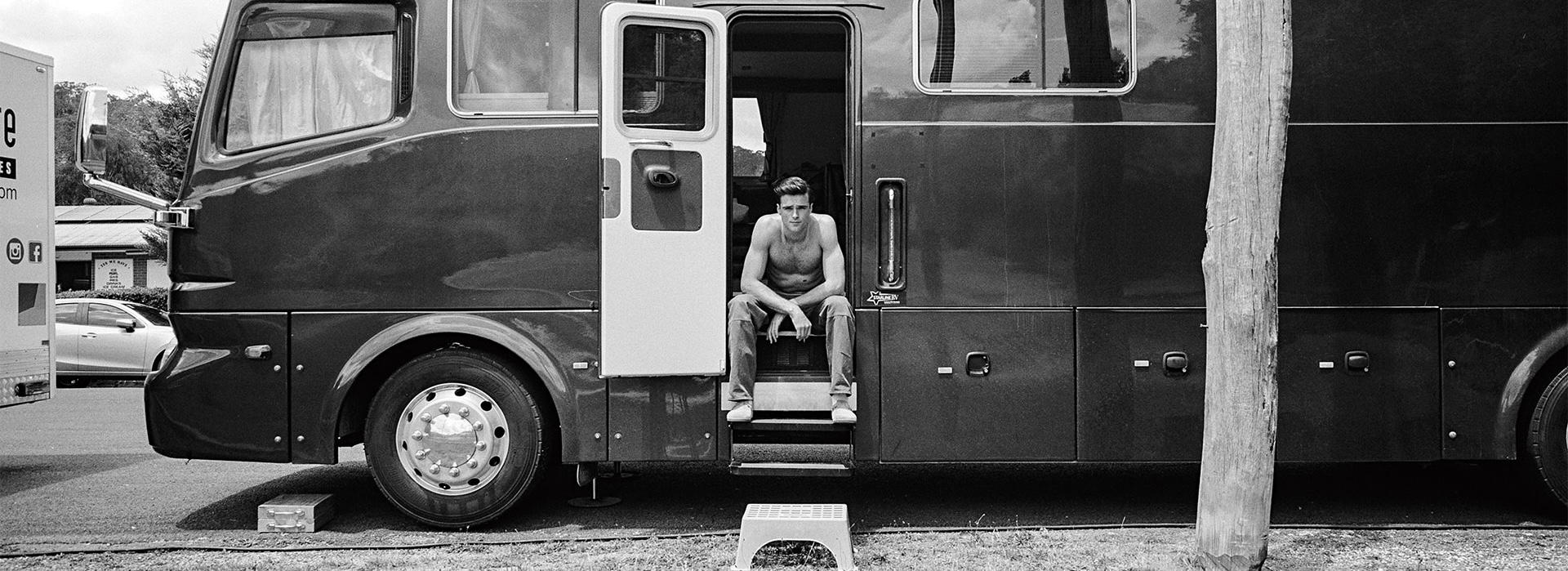 Black and white portrait of a shirtless man sitting in the doorway of a tour bus in the daytime, captured in a medium shot.