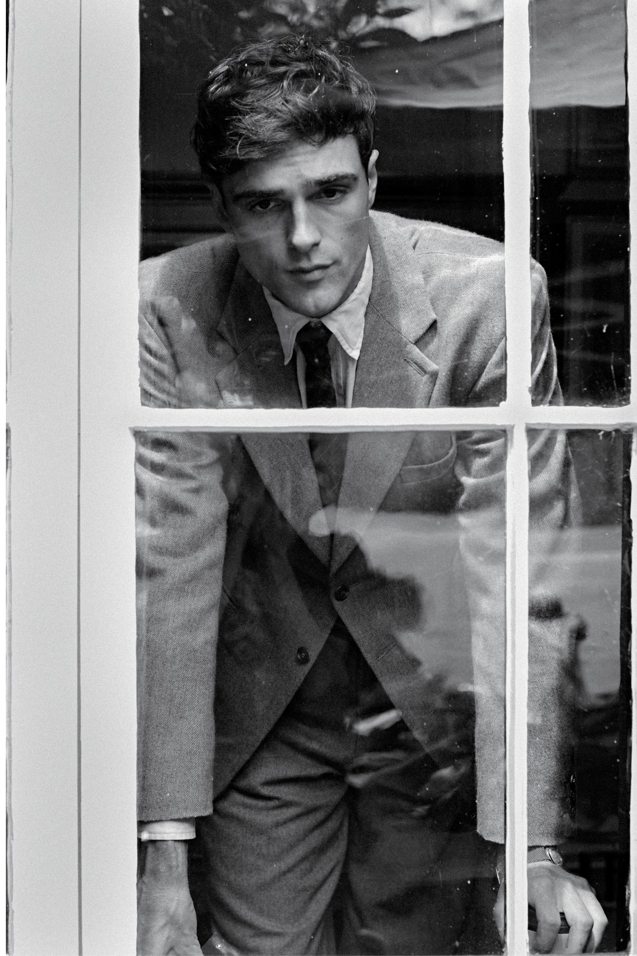 Jacob Elordi wearing a suit and tie looking through a multi-pane window in a black and white editorial portrait.
