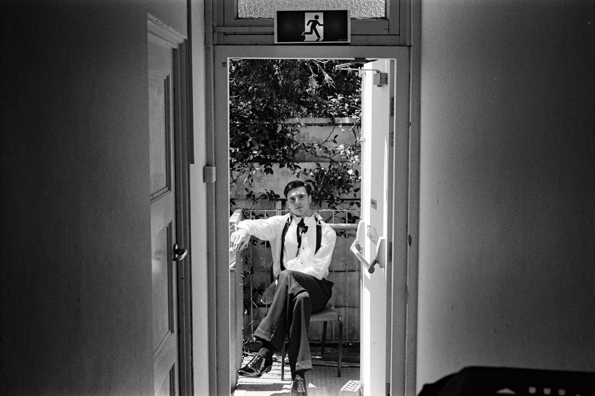 Jacob Elordi seated on a balcony outside an open doorway wearing a white shirt and dark trousers for Vogue Australia