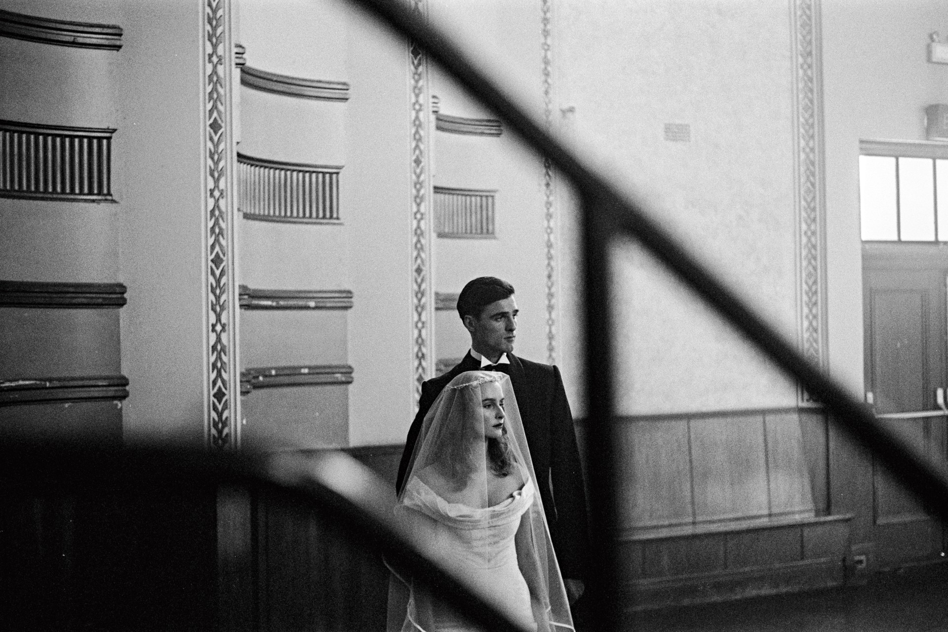 Black and white photograph of a bride and groom in front of paneled walls with decorative molding and window in a formal setting.
